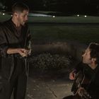 Zwei Männer draußen bei Nacht; einer spielt Gitarre, der andere hält eine Flasche. Im Hintergrund leuchten Lichter.