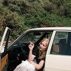 Eine Frau sitzt in einem offenen Auto und hält eine Zigarette. Im Hintergrund ist grüne Vegetation zu sehen.