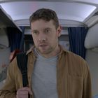 Ein Mann im Flugzeuggang trägt eine braune Jacke und Rucksack. Gepäckfächer sind offen, eine Person im Hintergrund.