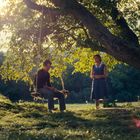 Keine sichtbaren Texte. Ein Mann auf einer Schaukel unter einem großen Baum, eine Frau steht daneben, Sonnenlicht strahlt durch die Blätter.