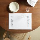 Weißes Tischset mit schwarzem Blumenmuster, weißer Teller, Wasserglas und Brötchen im Tuch auf Holztisch.