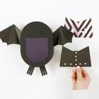 Papierfledermaus mit ausgebreiteten Flügeln, eine Hand hält ein Stück mit ausgestanzten Quadraten.