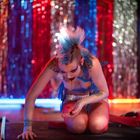 Es gibt keinen erkennbaren Text. Eine Person mit auffälliger Haarfrisur und Schminke kniet auf einer Bühne mit bunten Vorhängen.