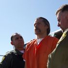 Zwei Polizisten in Uniform flankieren einen Mann in orangefarbenem Overall unter freiem Himmel.