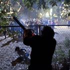 Ein Mann mit Kettensäge am Zaun, eine Person liegt am Boden. Im Hintergrund ein Rummelplatz mit Menschen.