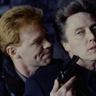 Zwei Männer in einer intensiven Szene, einer hält eine Pistole an den Kopf des anderen, im Hintergrund unscharfe dunkle Umgebung.