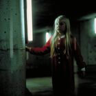 Ein Mädchen in rotem Pyjama steht in einem dunklen Raum vor einer Betonstütze, umgeben von leuchtenden Röhren.