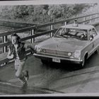 Ein Auto mit Kennzeichen "2-462" und einem rennenden Mann auf einer Brücke; der Fahrer schaut aus dem Fenster.