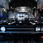 Ein schwarzes Auto in einem Flugzeug. Ein Mann steht daneben. Text: "Charger". Umgebung wirkt industriell und technisch.