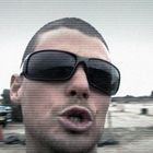 Ein Mann mit Sonnenbrille, nah aufgenommen; im Hintergrund ein blaues Auto auf einer staubigen Fläche.