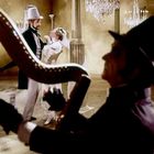 Ein Mann spielt Harfe, während ein Paar in eleganter Kleidung im Hintergrund in einem opulenten Raum tanzt.