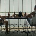 Zwei Frauen sitzen an einem Tisch mit Blick auf Wasser, trinken Bier, entspannen in einem rustikalen Innenhof mit Gitterstäben.