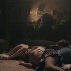 Dunkler Raum, eine Person sitzt am Tisch, im Vordergrund liegen mehrere Menschen am Boden.