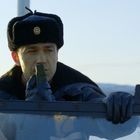 Ein Mann in Winterkleidung hält ein Funkgerät. Er trägt eine Pelzmütze mit einem Emblem darauf. Schneebedeckte Berge im Hintergrund.