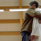 Zwei Personen umarmen sich in einem Raum mit Holzbalken und Leuchtstoffröhren an der Decke.