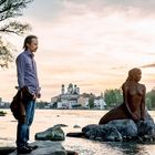 Ein Mann steht nachdenklich auf einem Felsen im Fluss, neben einer Meerjungfrauenstatue, mit einer Stadt im Hintergrund.