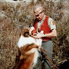Ein Collie springt an einem Mann in einer roten Weste hoch. Sie stehen in einer trockenen Wüstenlandschaft.