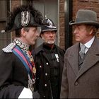 Zwei Männer in viktorianischer Kleidung stehen auf der Straße; einer trägt einen aufwendig verzierten Hut. Im Hintergrund ein Polizist.