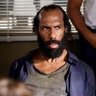 Ein Mann mit dichtem Bart und ernstem Gesicht sitzt in einem Raum. Jemand steht im Hintergrund mit einem weißen Hemd.