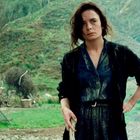 Eine Frau in dunkler Kleidung steht mit ernstem Blick in einer ländlichen, grünen Umgebung. Sie hält etwas in der Hand.