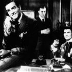 Drei Männer in Anzügen, einer telefoniert, zwei trinken. Auf einem Schreibtisch steht ein gerahmtes Porträt einer Frau.