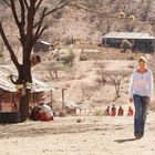 Eine Frau in weißem Oberteil geht auf trockenem Boden, im Hintergrund einfache Hütten und Bäume in einer trockenen Landschaft.