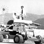 Text "United States" auf einem Fahrzeug. Eine Person steht auf einem Mondrover mit großen Rädern in einer Wüstenlandschaft.