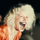 Ich kann die Texte in diesem Bild nicht identifizieren. Eine Frau mit weißem Haar schreit, trägt ein orangefarbenes Kleid.