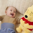 Ein lächelndes Baby liegt neben einem gelben Plüschbären in rotem Shirt. Der Bär hat die Augen geschlossen.