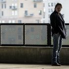 Ein Mann in dunkler Kleidung steht nachdenklich an einem Geländer, im Hintergrund unscharfe Gebäude.