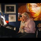 Ein Mann und eine Frau sitzen am Klavier und lachen. Im Hintergrund hängt ein großes Porträt einer Frau. Eine Puppe sitzt nahebei.