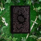 Verziertes schwarzes Buch auf Gras, umgeben von Pflanzen mit Dolch. Auf dem Buch sind florale Muster und Symbole geprägt.
