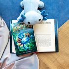 "THE IMMORTALIS: DAWN OF THE AXOLOTL" von Kit Brooks liegt geöffnet. Ein blauer Plüsch-Axolotl liegt darauf.