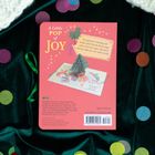 Text: "A Little POP of JOY" und "This gift book is bursting with holiday spirit..." Illustration: Weihnachtsbaum-Popup auf rotem Hintergrund.