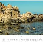 "Cala Vadella", "Casares Edition", Monatsübersicht Juni. Felsige Küste mit klarem Wasser; im Hintergrund eine alte Steinhütte.