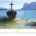 "Montsee" und "6". Ein See mit Bergen im Hintergrund, ein Felsen mit zwei Kreuzen steht im klaren Wasser.