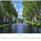 Text: Kö-Graben. Unter einer Baumallee verläuft ein ruhiger Fluss, umgeben von grünen Wiesen und historischen Gebäuden.