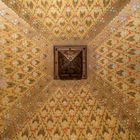 Eine goldene Decke mit geometrischem Muster und Blütenverzierungen, in der Mitte ein geschnitztes Holzpaneel.