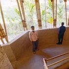 Zwei Personen stehen in einem Gebäude mit Bambusstangen und Strohdach, umgeben von Palmen.