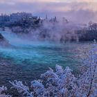 Rheinfall nahe Schaffhausen bei Winterlandschaft; Schloss im Hintergrund, gefrorene Bäume, Wasserfall und Brücke sichtbar.