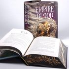 "EMPIRE OF BLOOD: WE WHO WILL DIE." Aufgeschlagenes Buch davor, Cover mit goldener geflügelter Kreatur in Lorbeerkranz.