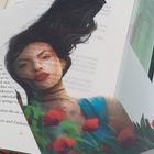 Ein Buch mit Textpassagen. Darauf ein Transparent mit einer Frau und roten Blumen. Ihr Haar weht, sie trägt Blau.