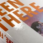 Der Text lautet „BEEN THERE,“ in großen orangefarbenen Buchstaben. Darunter sind gemalte Figuren zu erkennen.
