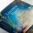 Text: "Lucinda Riley. Die Frauen von Ballymore. Roman." Buchcover mit Blumenmotiv in Blau und Gold.