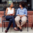 "Die sanfte Seite von New York." Zwei Frauen sitzen auf einer Bank vor einem Café und unterhalten sich. Links neben ihnen ist ein Schild.