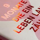 "9 MONATE DIE EIN LEBEN LANG ZÄHLEN" in glänzenden Buchstaben auf rosa Hintergrund, Herzform im Design.