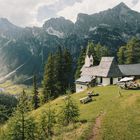 Eine weiß verputzte Kirche mit spitzem Turm steht auf einem grünen Hang vor einer beeindruckenden Bergkulisse.