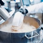 Eine Person in Schutzkleidung arbeitet in einer Industrieanlage. Pulver wird in einem großen, runden Behälter abgesaugt.