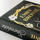 „EMILY BRONTË“ und „Sturmhöhe“ in goldener und weißer Schrift, umgeben von goldenen Ranken und weißen Blumen.