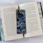 Ein geöffnetes Buch zeigt Text. Ein blauer Lesezeichen mit floralen Mustern liegt darauf.